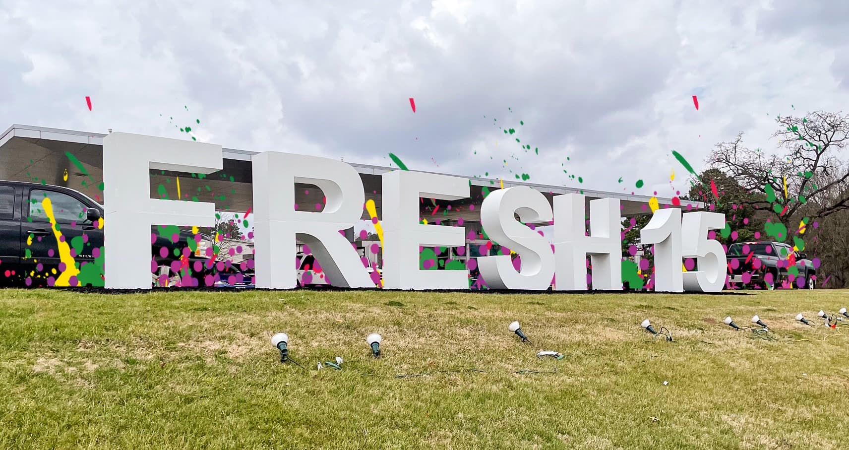 A large, colorful "FRESH 15" sign sits on a grassy area under a cloudy sky.