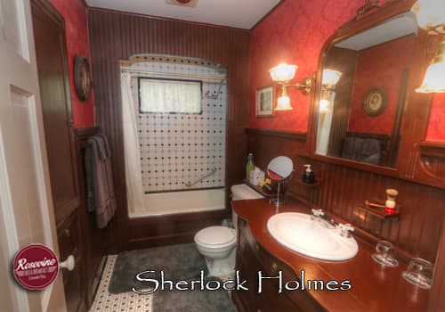 A private bed & breakfast cottage bathroom with red wallpaper and brown wainscotting. White subwaytile tub and shower combo, commode and brown wood sink