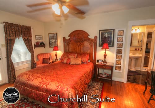 A private extended stay room with white walls, wood floors, brown highback bed with red and gold duvet. There is also a wood desk in the room