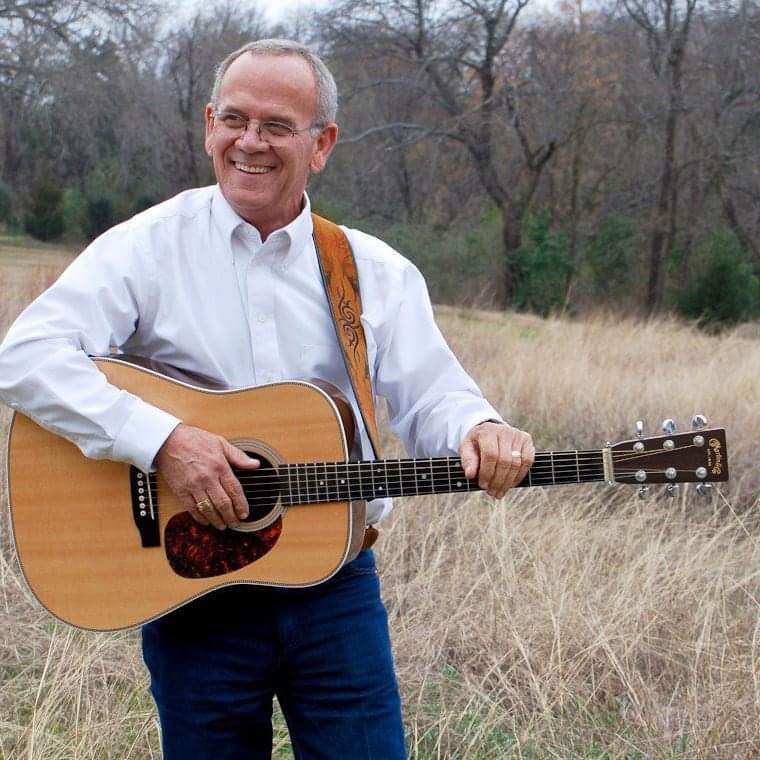 A smiling man in a white shirt plays acoustic guitar in a grassy field.