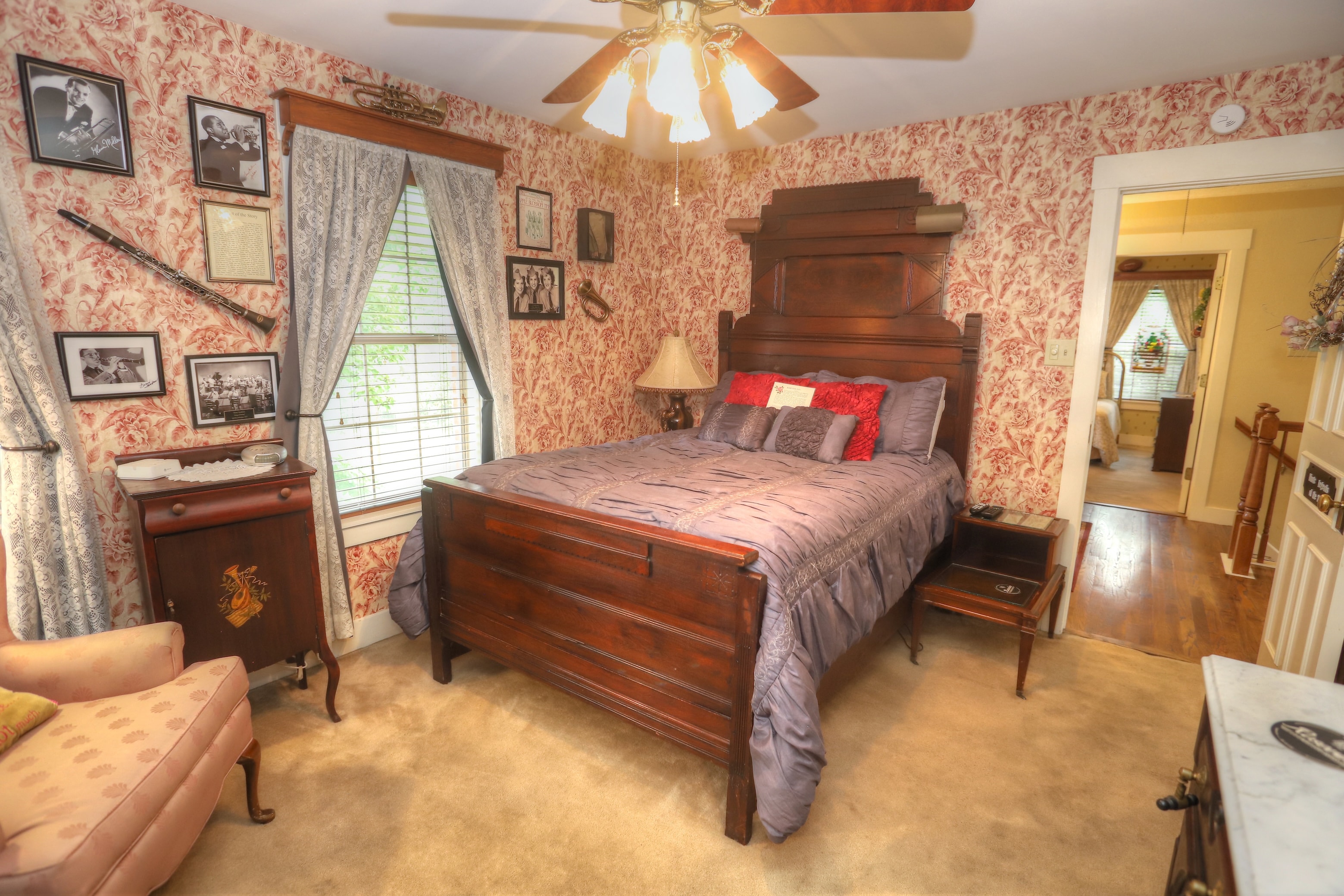 An upstairs bed and breakfast room with red and creme wall paper, a dark brown highback bed with a blue duvet. Also in the room is a pink fabric chair and brown ceiling fan