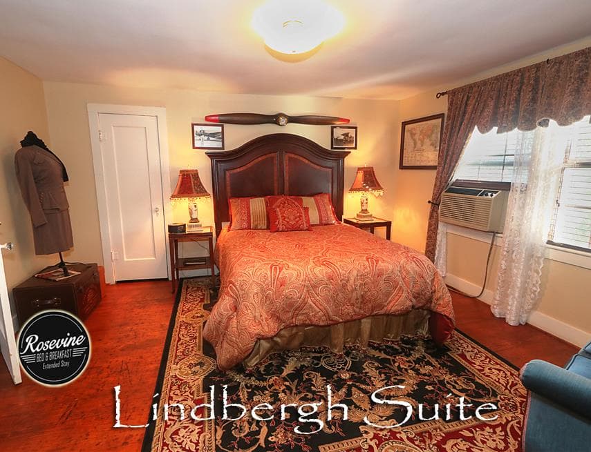 The upstairs Lindbergh Suite Bedroom featurers a wood headboard bed with red and gold duvey. Also in the room is a window unit, blue fabric chair and closet