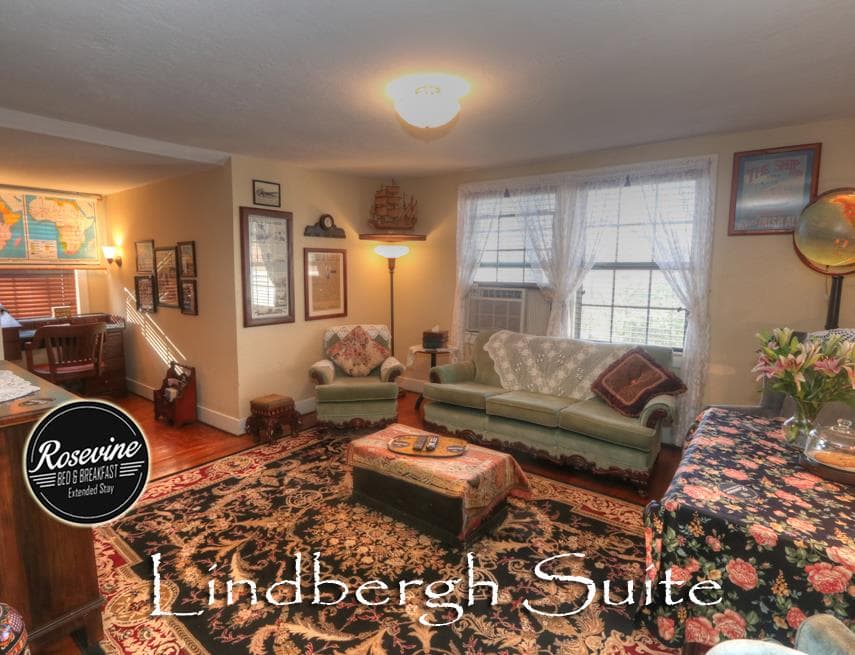 The upstairs Lindbergh Suite Living Room features creme walls, wood floors, black floral rug, green fabric soft, a wood desk and tv