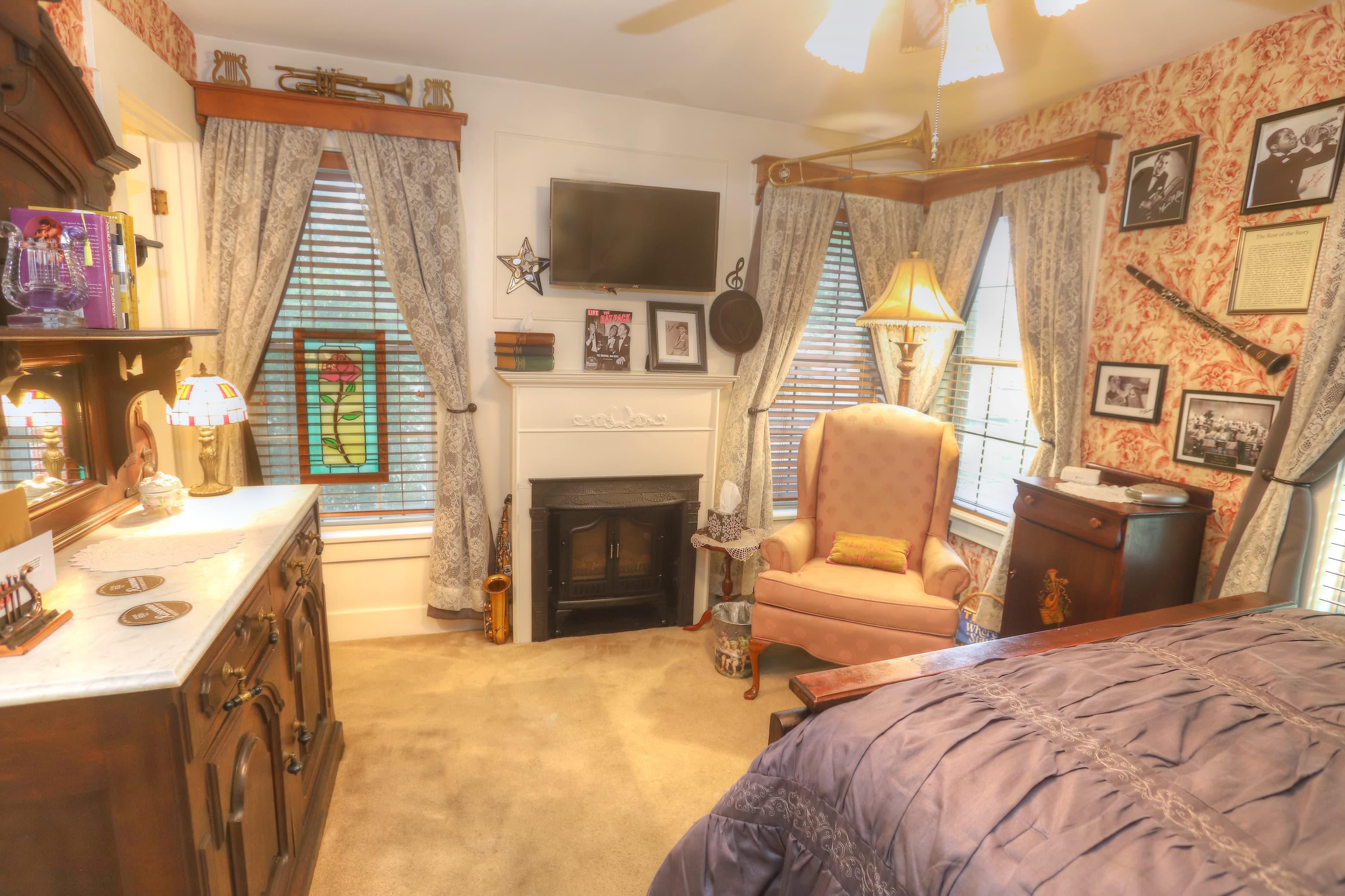 An upstairs bed and breakfast room with red and creme wall paper, a dark brown highback bed with a blue duvet. Also in the room is a pink fabric chair and brown ceiling fan