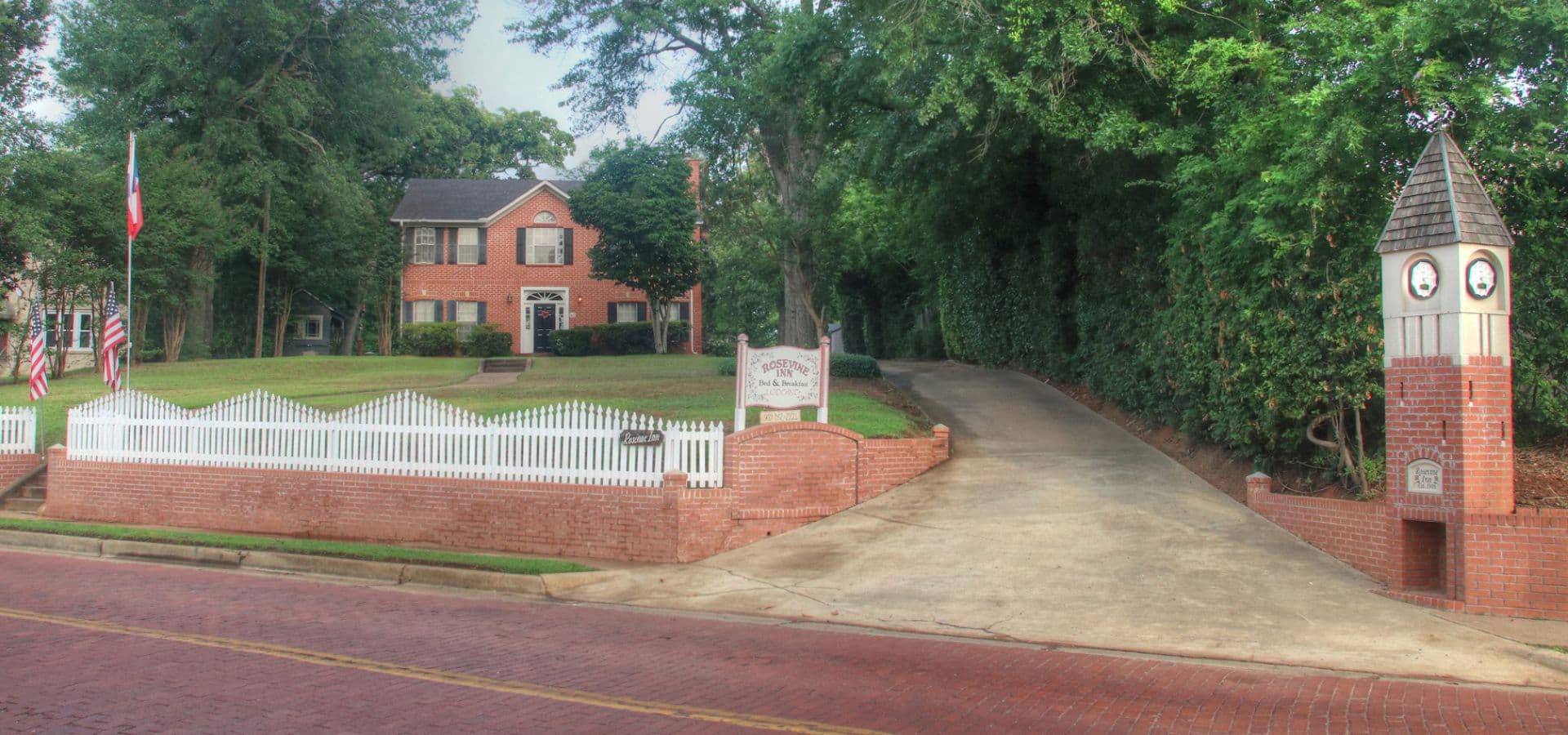 A brick house with a white picket fence and a clock tower near the driveway, surrounded by trees.