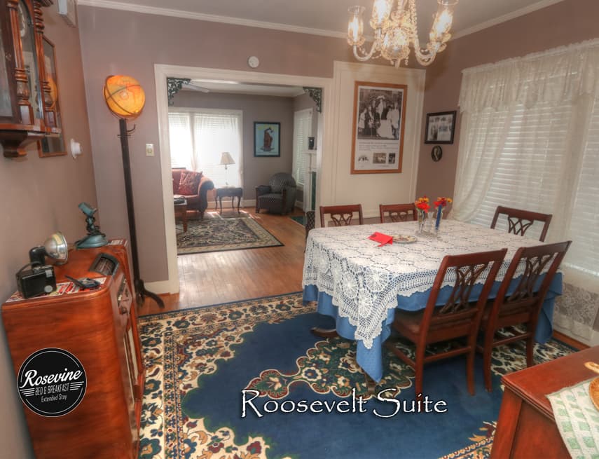 Roosevelt dining room with wood table that has a blue table cloth and lace overlay. Four wood chairs, blue rug, lamp in the shape of a globe and small chandelier
