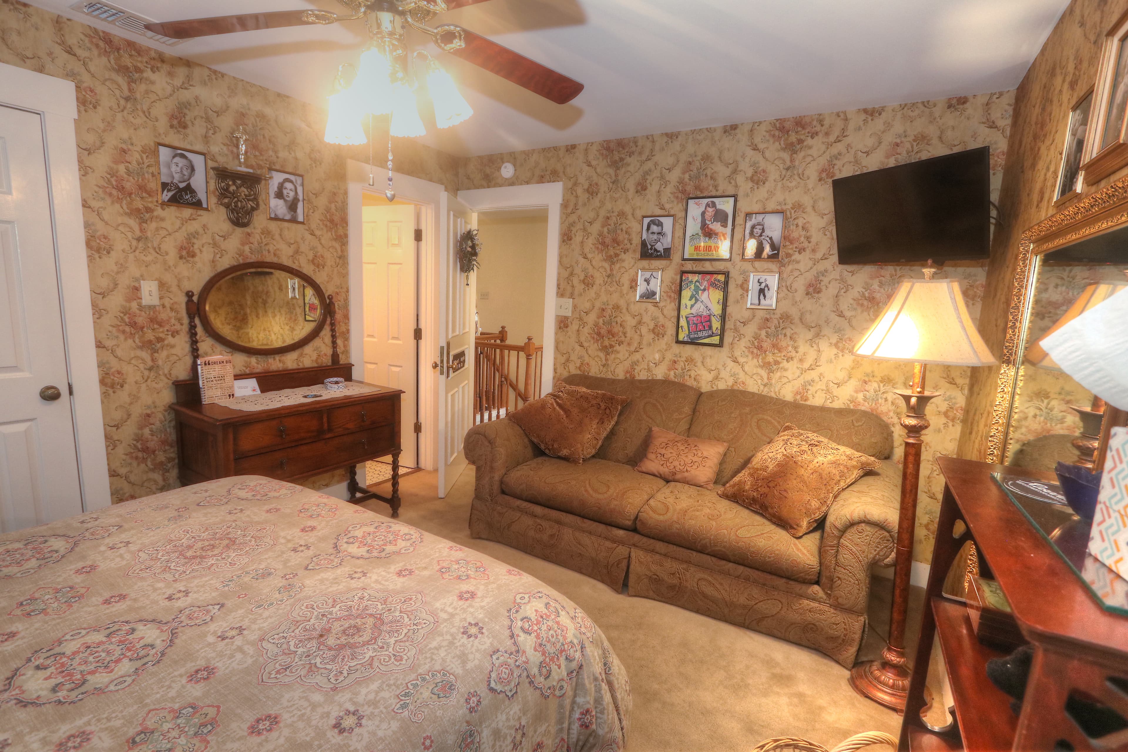 An upstairs bed and breakfast room with brown and floral wall paper, a decorative white metal bed with a red duvet. Also in the room is a brown loveseat and brown ceiling fan