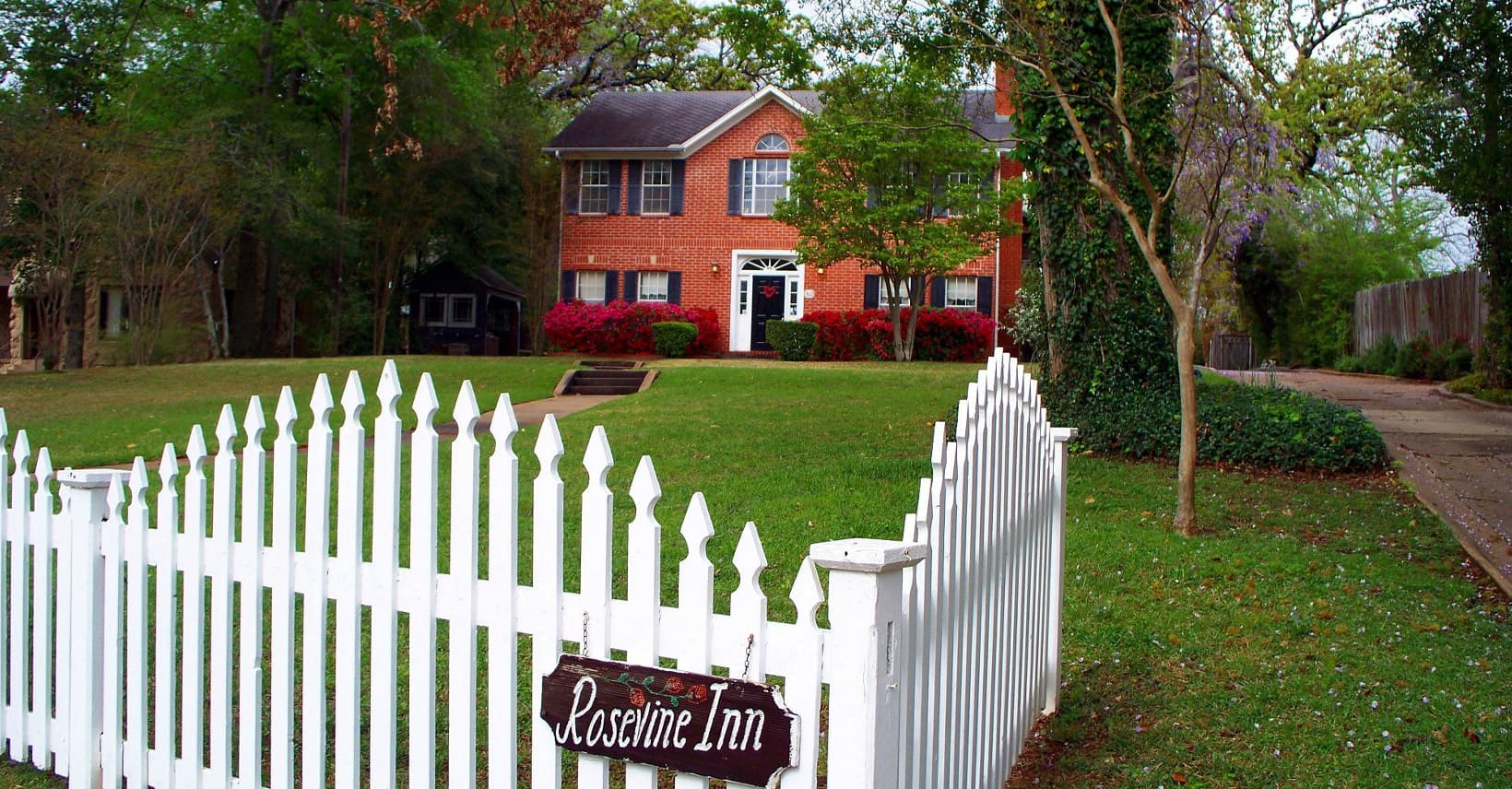 A brick house with a white picket fence and a sign reading "Rosewine Inn" in a green yard. A brick house with a white picket fence and a sign reading "Rosewine Inn" in a green yard.