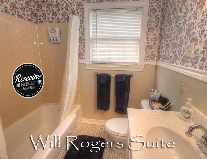 Will Rogers Suites bathroom with creme walls, floral wall paper, tub shower combo, commode and sink