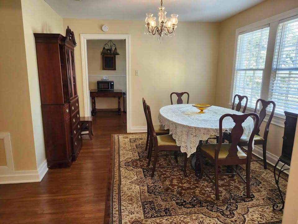A dining room featuring a round table with a lace tablecloth, surrounded by wooden chairs, an elegant chandelier, and light-colored walls.