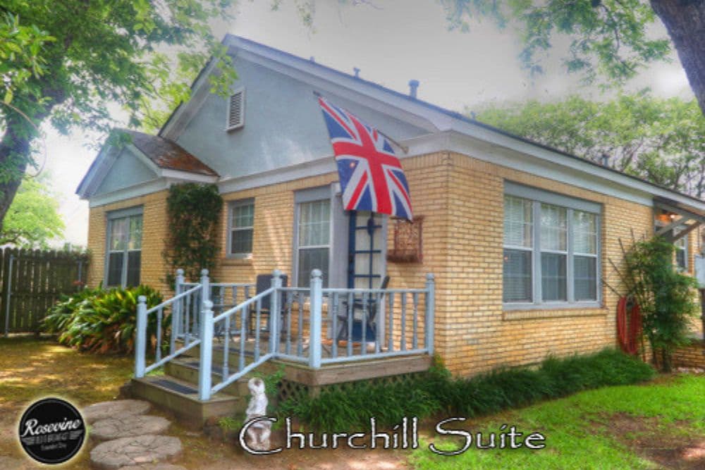 A charming yellow-brick cottage featuring a British flag, surrounded by greenery, with a sign reading "Churchill Suite."