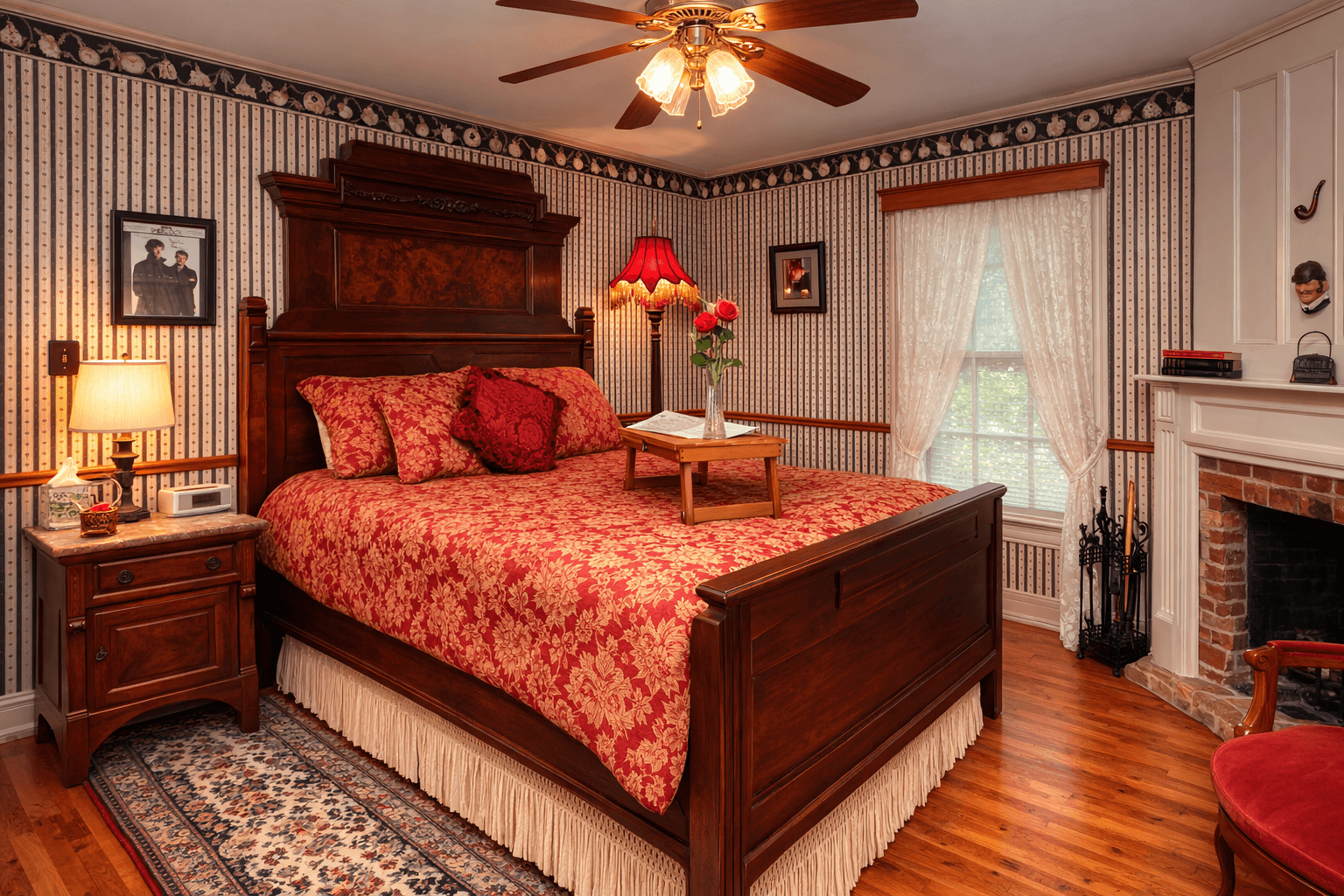 A private bed & breakfast cottage bedroom with green and white striped wallpaper, a dark brown highback bed with red duvet. Also in the room is a pink fabric chair, mock fireplace and brown ceiling fan