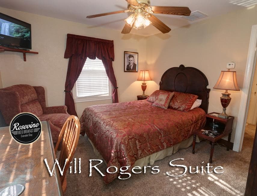 An upstairs bed and breakfast suite with creme walls, light brown carpet, a bed with brown headboard and red/gold duvet. Also in the room is a decorative wood shelf/desk combo