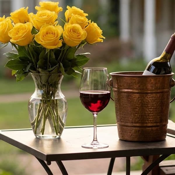 A glass of red wine next to a bouquet of yellow roses and a wine bucket on a table outdoors.