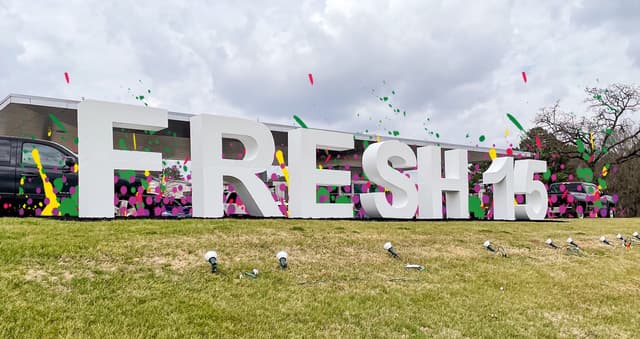 white letters in a grassy setting says Fresh15-has colorful flowers behind the letters