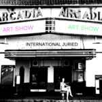 Arcadia Marquee with art show on one side in pink on the other side in green. A mannequin is seated out front on the sidewalk. Words above her head say International Juried
