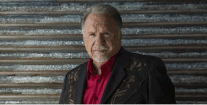 photo of Gene Watson in a red shirt with a black Jacket standing in front of a metal wall