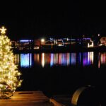 Christmas tree decorated with white lights on a lake. It is located in the forground on te left. Across the lake are many different colored Christmas lights reflecting in the lake