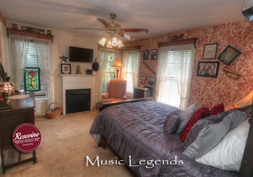 An upstairs bed and breakfast room with red and creme wall paper, a dark brown highback bed with a blue duvet. Also in the room is a pink fabric chair and brown ceiling fan