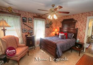 An upstairs bed and breakfast room with red and creme wall paper, a dark brown highback bed with a blue duvet. Also in the room is a pink fabric chair and brown ceiling fan