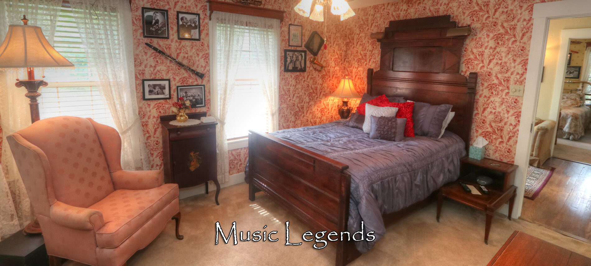 An upstairs bed and breakfast room with red and creme wall paper, a dark brown highback bed with a blue duvet. Also in the room is a pink fabric chair and brown ceiling fan