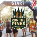 mage of a staring gate for a run with several people in running clothes in the middle is a log with pine trees on top and under that the words Run the Pines Trail Series