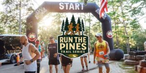 Image of a staring gate for a run with several people in running clothes in the middle is a log with pine trees on top and under that the words Run the Pines Trail Series