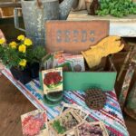 Plethora of garden items spread out on a quilt on top of a wooden table . Sign in the background says seeds 