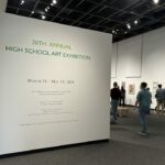 Sign in the foreground in green lettering saying 20th Annual Hiugh School Art Exhibition. people are milling around looking at the art.