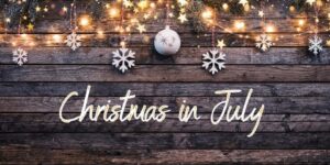 Chirstmas ornaments and lights on a wooden surface with the words Christmas in July under them
