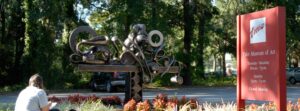 Picture of the driveway in front of Tyler Museum Of art-person sitting on a bench behind a metal modern art sculpture -to the right is a red sign that says TMA Tyler Museum of Art