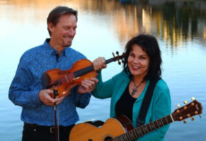 Picture of Gordon and Christy McLeod standing infront of water. He is holding a violin and she is holding a guitar