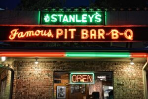 Neon sign on a brick building green letters says Stanleys under that is orange leters saying Famous Pit Bar-B-Q the a strip of neon red and green-under that in the window os a red open sign with a green border