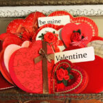 serveral valentine heart boxes that may have chocolates in them there are a few red silk roses attached and the words be mine on one small sign at top center and velos that is a sign that says Valentine