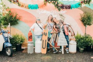 3 ladies smiling with pink pastels backfround. The words Vintage Market is written above thier heads in white lettering. Several random items surroundf the ladies that they are shopping for