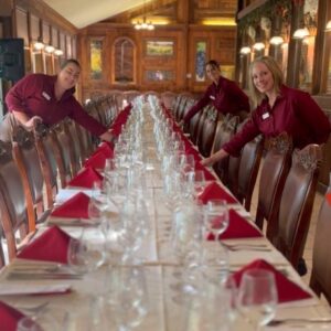 long table set elegantly with white table and red napkins. Lots of wine glasses. Three staff members are shown setting table